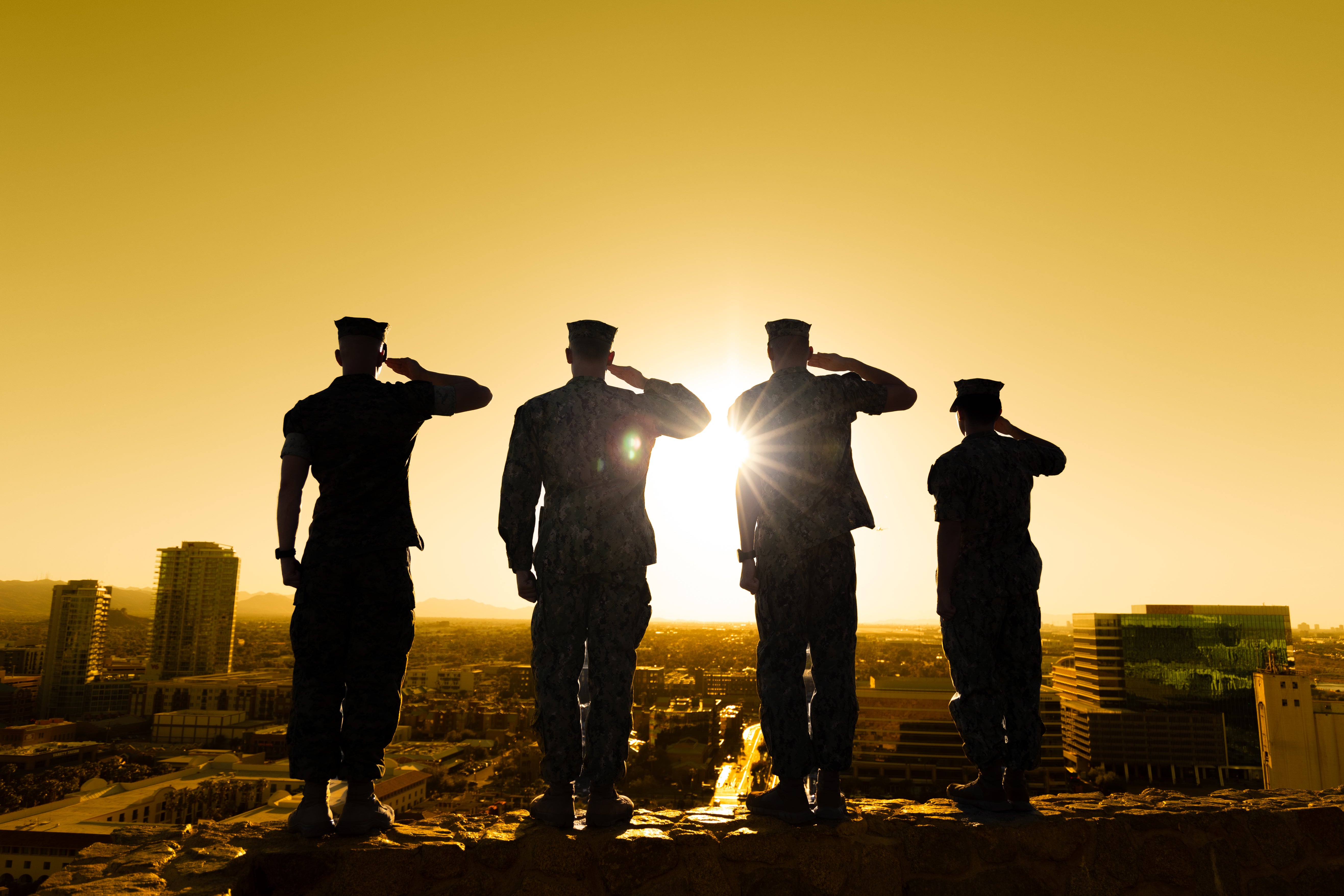 Four military personnel silhouetted against a golden sky while saluting, with a cityscape in the background.