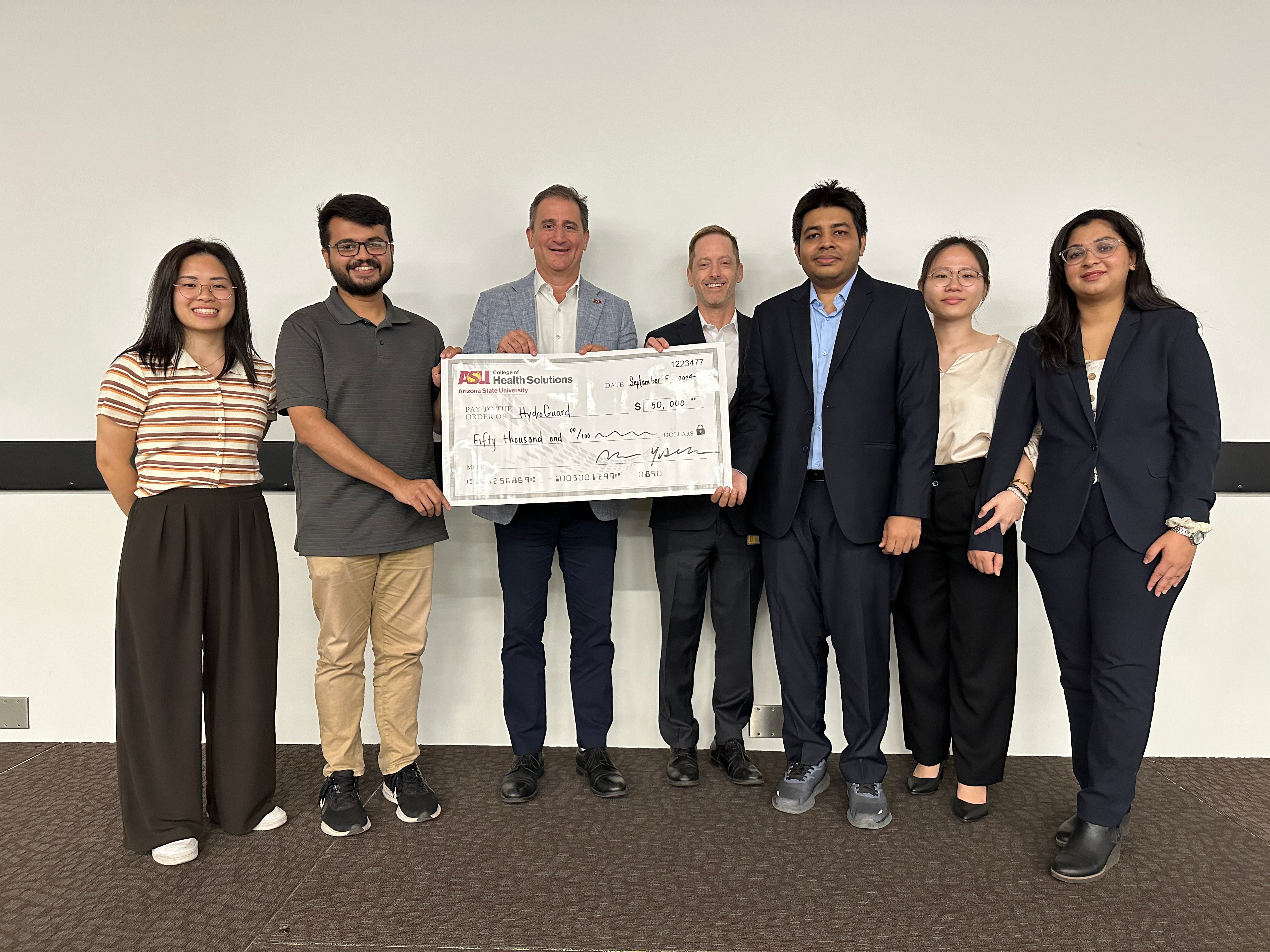 Group of seven people, two holding a large ceremonial check, posing in business attire.