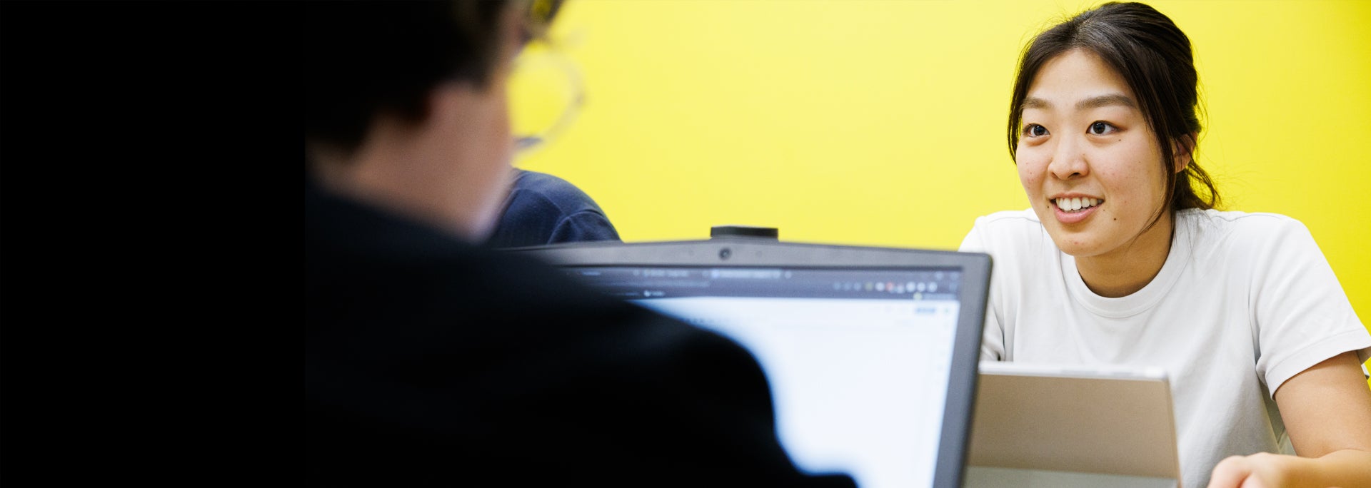 Two people talking in front of a bright yellow background, with one person blurred in the foreground.