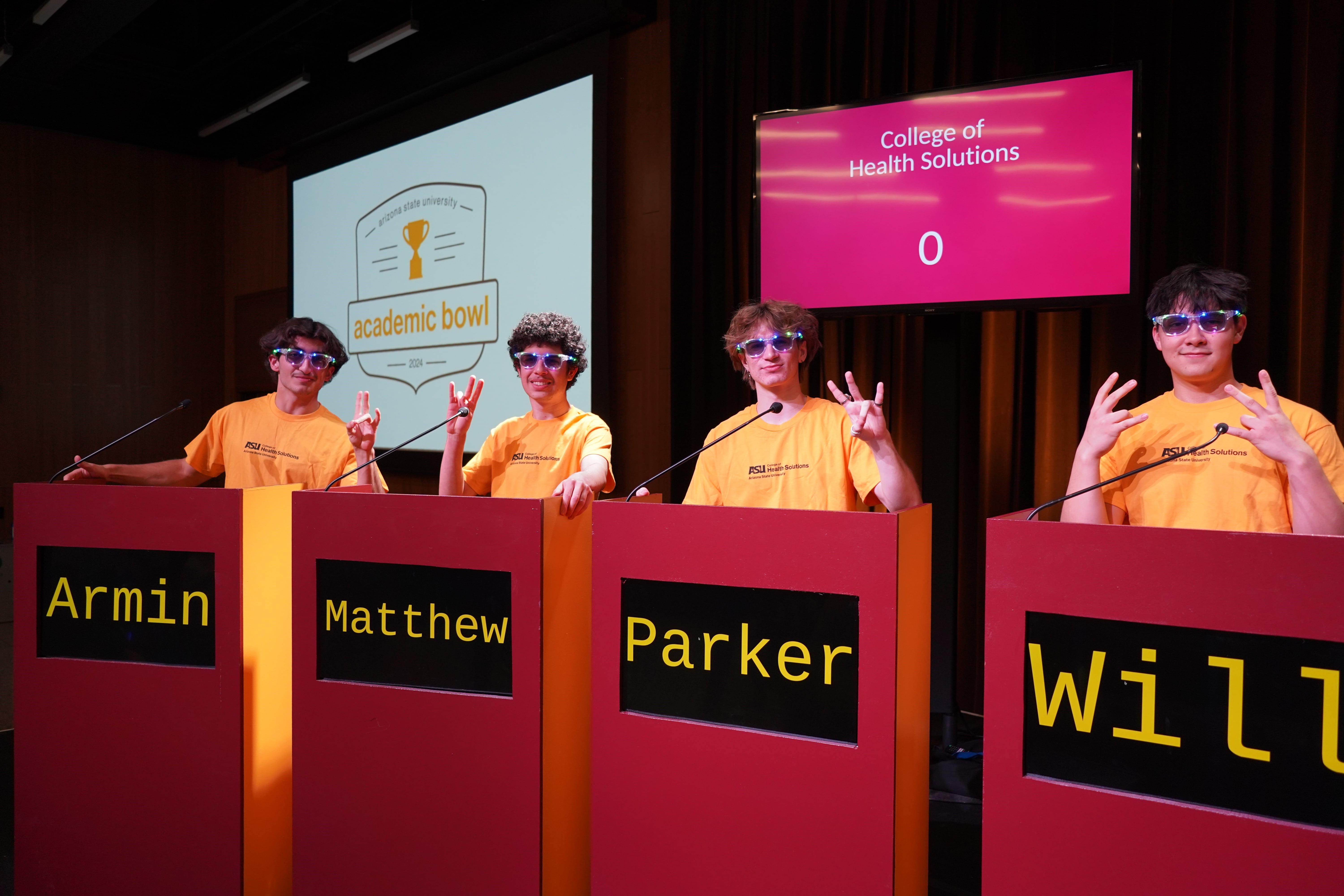 Four participants in yellow ASU Health Solutions shirts and colorful sunglasses stand behind podiums labeled Armin, Matthew, Parker, and Will, posing enthusiastically at the 2024 Arizona State University Academic Bowl.