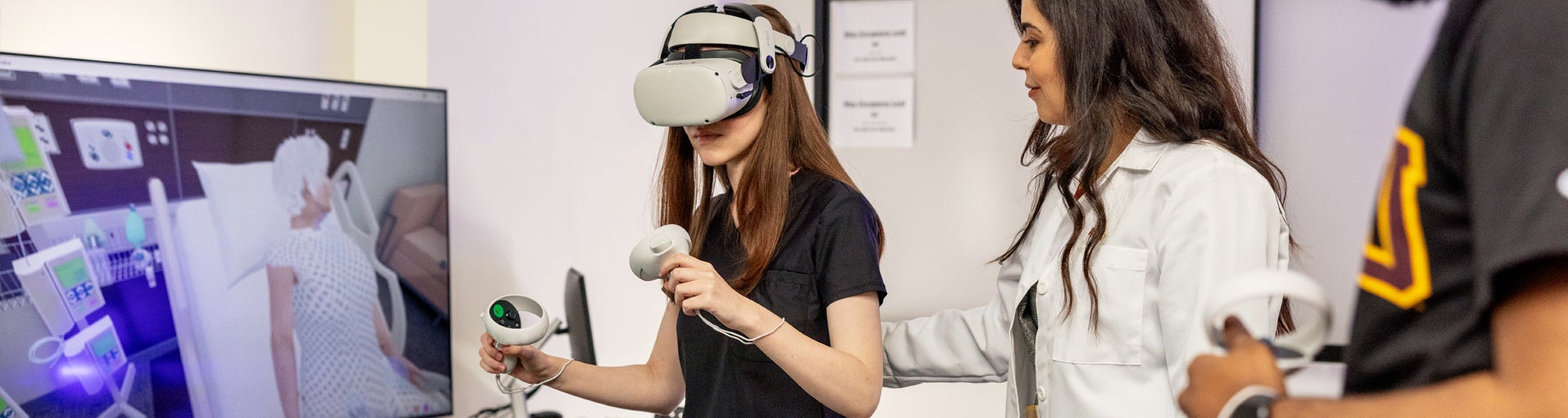 A woman uses a VR headset and controllers, assisted by another woman in a lab coat, with a medical simulation on a screen in the background.