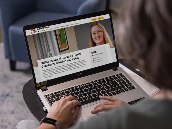 A person sitting at a table using a laptop displaying ASU Online's health care master's program page.